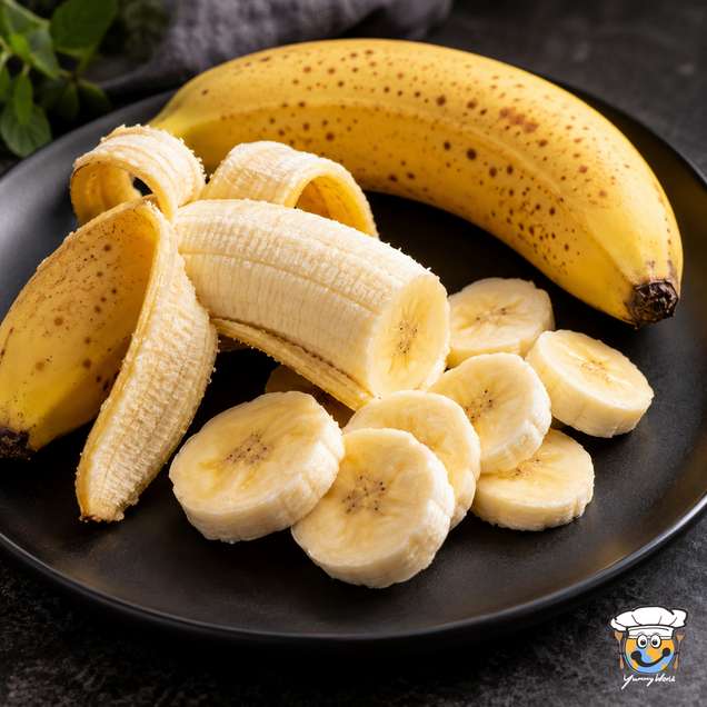 Eine reife ganze Banane neben einer geschälten Banane und dicken Bananenscheiben auf einem schwarzen Teller.