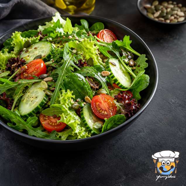 Gemischter grüner Salat mit Gurkenscheiben, Kirschtomaten und Kernen in einer schwarzen Schale.